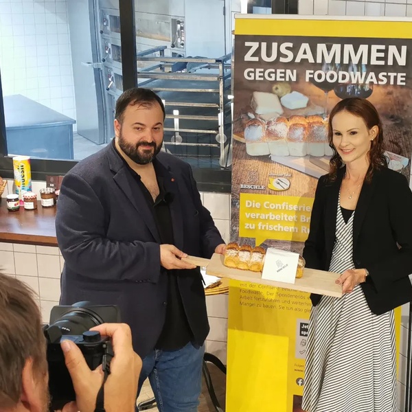 Zwei Personen stehen vor einem Banner mit der Aufschrift 'ZUSAMMEN GEGEN FOODWASTE' und halten gemeinsam ein Brett mit Brot.