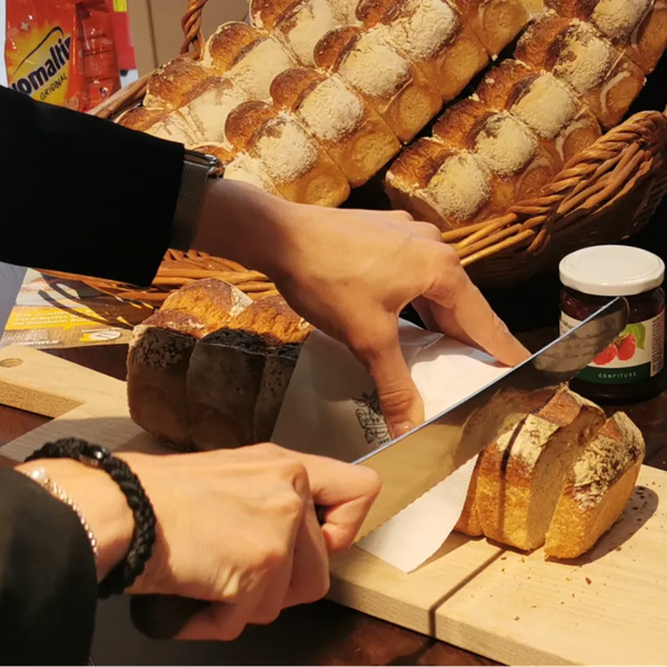 Zwei Hände schneiden ein helles, längliches Brot auf einem Holzbrett, im Hintergrund mehrere Brote in einem Korb und Gläser mit Aufstrich.