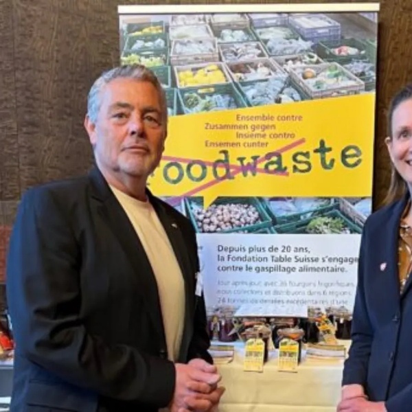 Zwei Personen stehen vor einem Stand mit Lebensmitteln und einem gelben Banner mit dem Schriftzug 'foodwaste'.