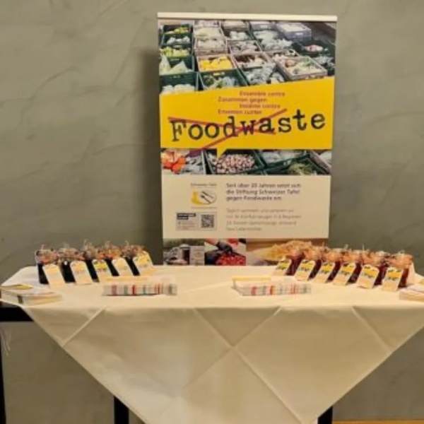 Tisch mit Informationsmaterial und kleinen Gläsern vor Plakat mit dem Schriftzug 'Foodwaste' in einem Raum mit Holztür und Fenster.