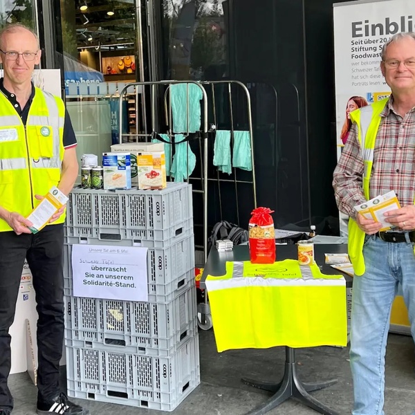 Zwei Personen mit gelben Sicherheitswesten stehen neben einem Tisch mit gelber Weste und Informationsmaterial vor einem Laden.