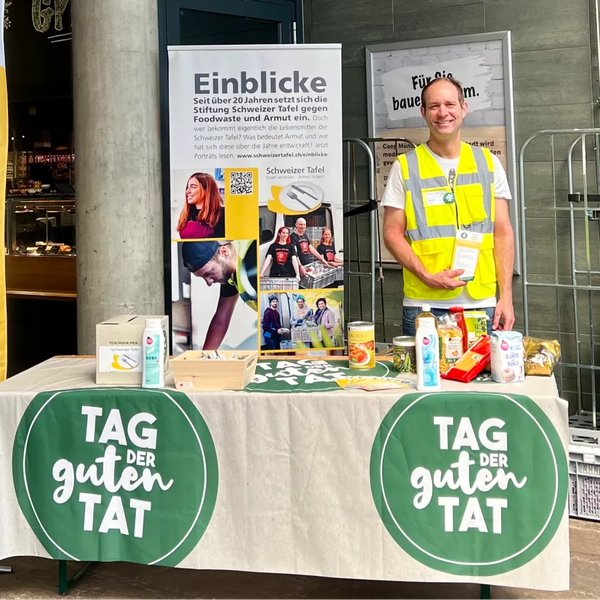 Mann mit gelber Warnweste steht an Tisch mit Tischtuch 'Tag der guten Tat' und verschiedenen Lebensmitteln vor einem Coop.