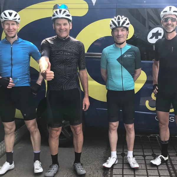 Vier Radfahrer in Sportkleidung und Helmen stehen vor einem dunkelblauen Fahrzeug mit gelbem Schriftzug.