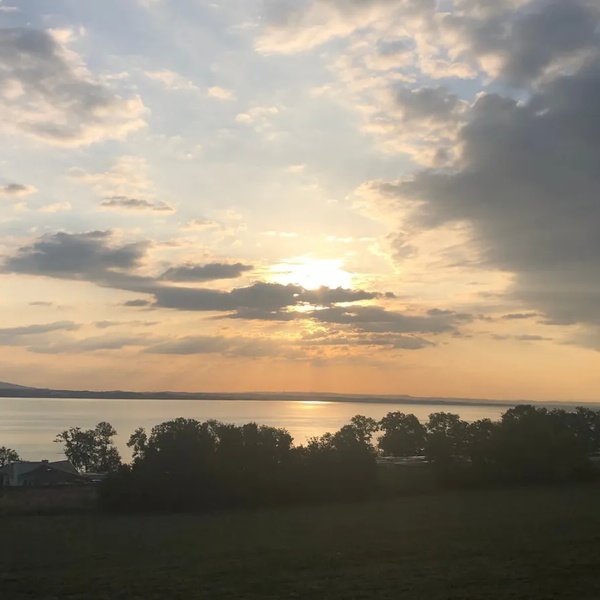 Sonnenuntergang über einem See mit bewölktem Himmel und Bäumen im Vordergrund.