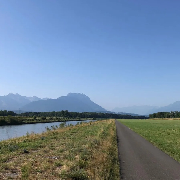 Asphaltierter Rad- und Fussweg neben einem Fluss mit Wiesen und Bergen im Hintergrund unter klarem Himmel.