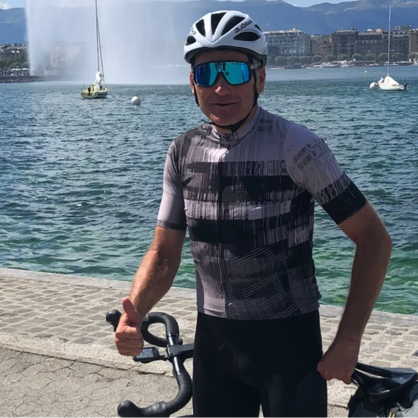 Radfahrer in schwarzem und grauem Trikot mit wessem Helm steht am Ufer eines Sees mit Springbrunnen und Segelbooten im Hintergrund.