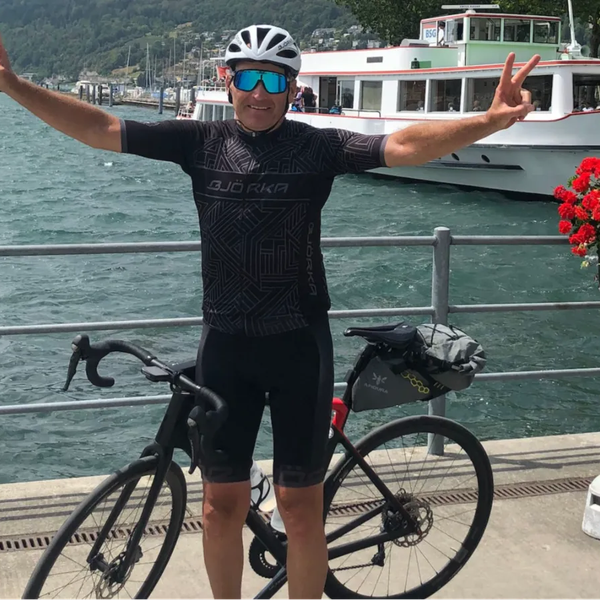 Mann in schwarzer Radbekleidung steht mit ausgestreckten Armen vor einem schwarzen Rennrad am Seeufer, im Hintergrund ein Boot und rote Blumen.