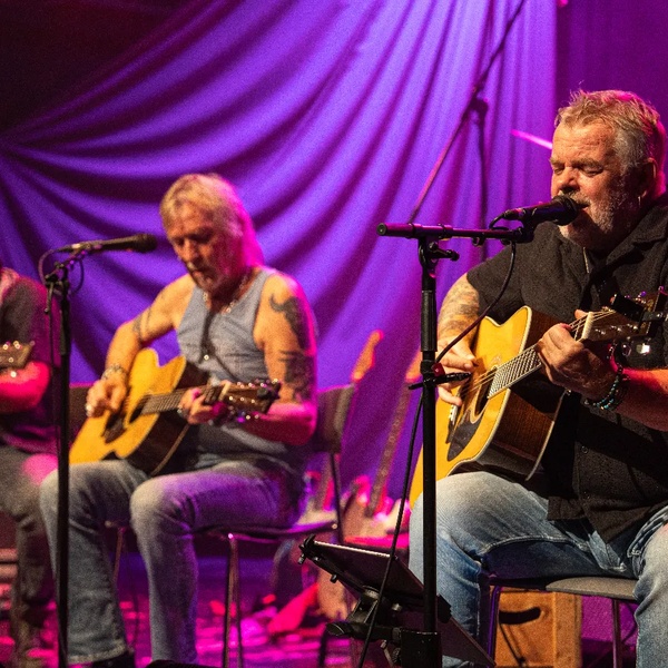 Die Band MGM spielt akustische Gitarren auf einer Bühne mit lila Hintergrundvorhang.