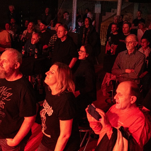 Gruppe von Menschen in einem Konzertraum mit roter Beleuchtung, einige tragen T-Shirts mit Aufdrucken, andere halten Getränke oder Handys.