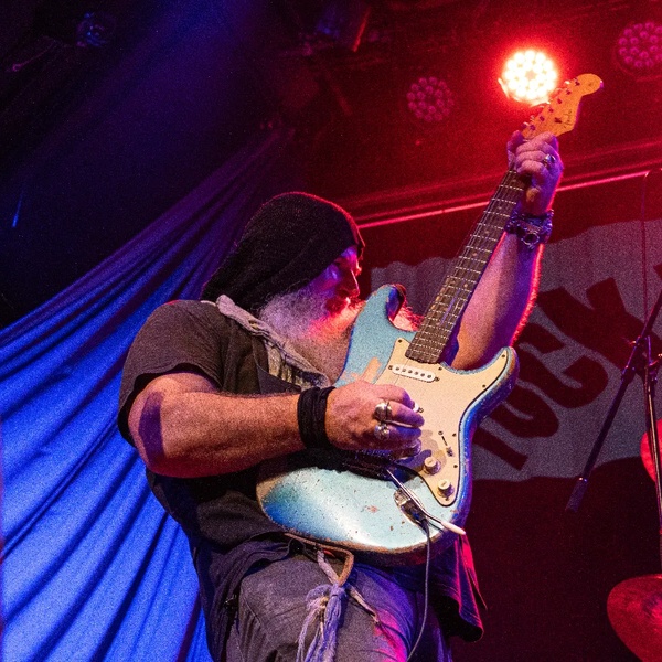 Gitarrist der Band Bluedög mit schwarzer Mütze und grauem Bart spielt E-Gitarre auf Bühne mit roter und blauer Beleuchtung.