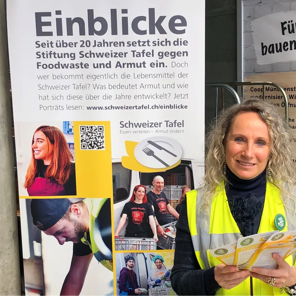 Frau mit gelber Warnweste hält Flyer vor einem Informationsplakat der Stiftung Schweizer Tafel.