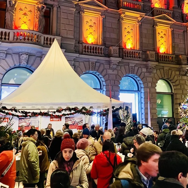 Menschenmenge vor zwei weissen Zelten und einem grossen, geschmückten Weihnachtsbaum vor einem beleuchteten historischen Gebäude.