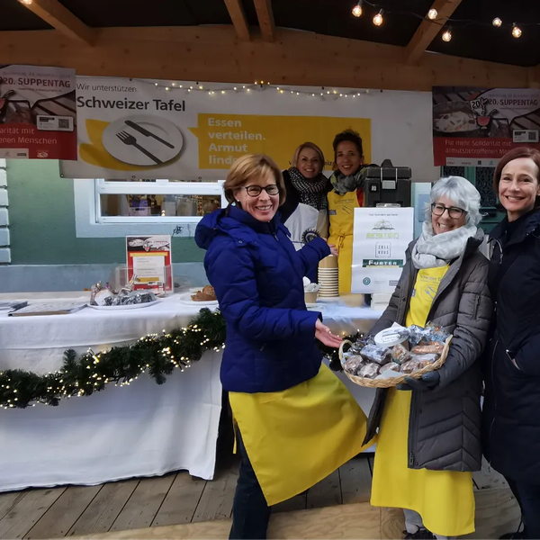Drei Personen stehen vor einem festlich geschmückten Stand mit Bannern der Schweizer Tafel, zwei tragen gelbe Schürzen, eine hält ein Tablett mit verpackten Lebensmitteln.