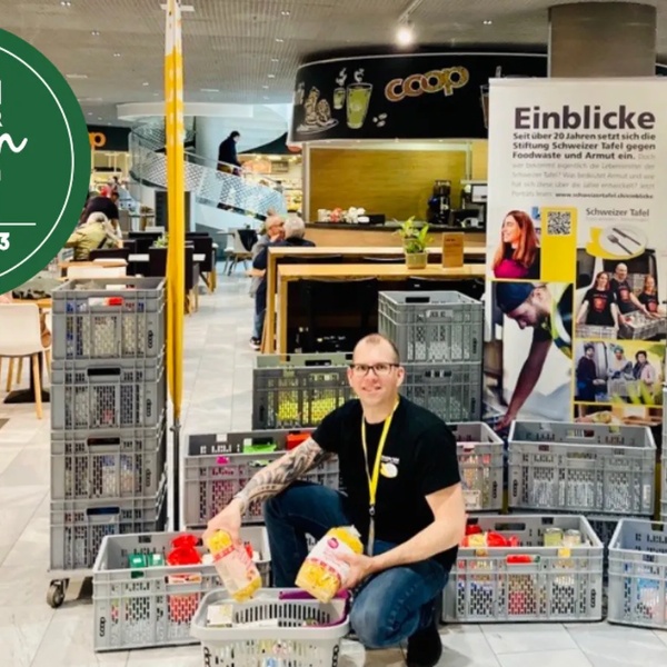 Mann mit schwarzem T-Shirt und gelbem Namensschild kniet vor Stapel aus grauen Kisten mit Lebensmitteln in einem Supermarkt.