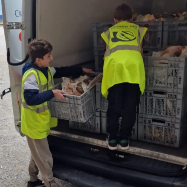 Zwei Kinder mit gelben Schweizer Tafel Warnwesten laden Kisten mit Nahrungsmittel aus einem weissen Lieferwagen aus.