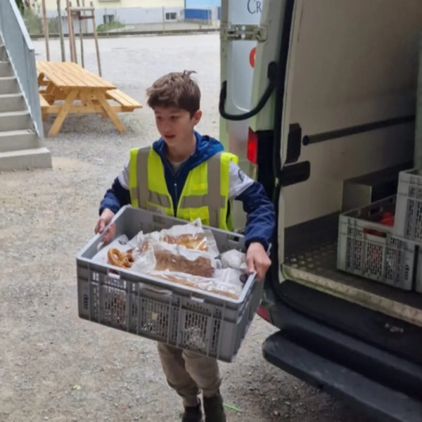 Zwei Kinder mit gelben Schweizer Tafel Warnwesten laden Kisten mit Nahrungsmittel aus einem weissen Lieferwagen aus.